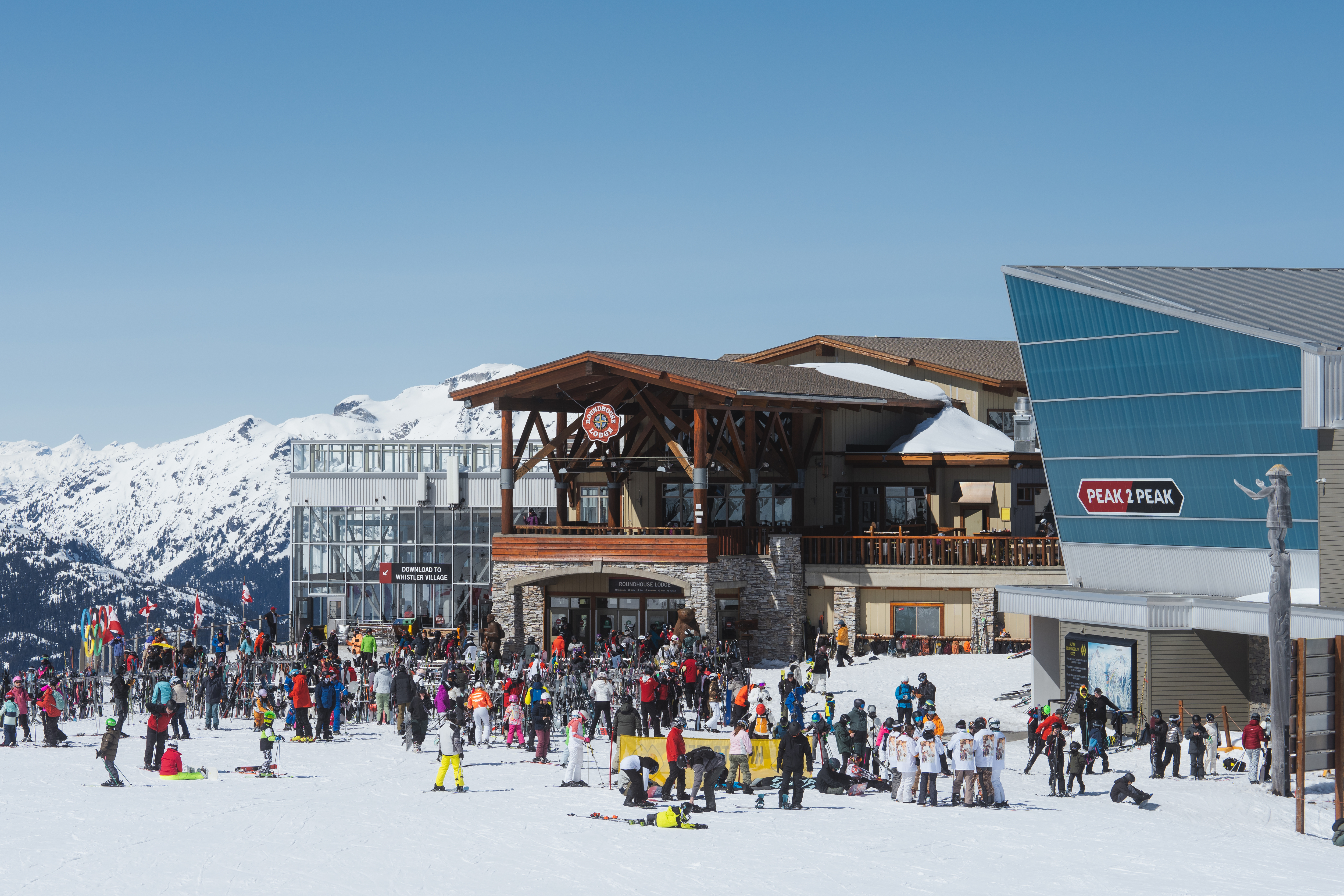 Ski lodge building with a wide, ground‑level entrance and open double doors. People in ski gear gather outside on a cleared, level entry area with railing and spacious pathways leading into the lodge.