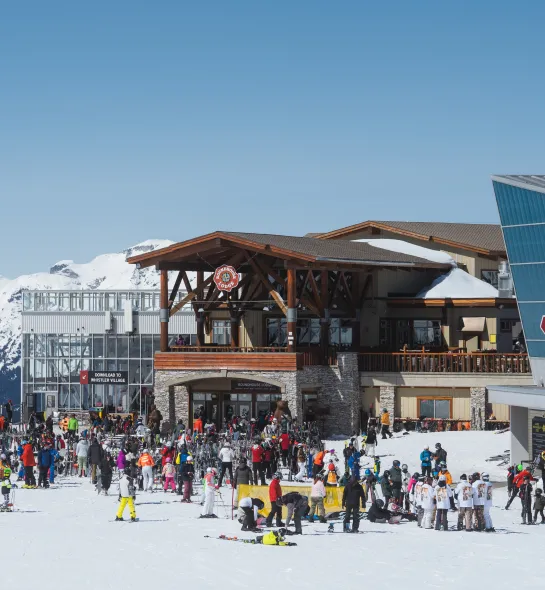 Un b&acirc;timent de la station de ski dot&eacute; d&rsquo;une large entr&eacute;e &agrave; doubles portes au niveau du sol. Des personnes en tenue de ski se rassemblent &agrave; l&rsquo;ext&eacute;rieur, sur un espace d&eacute;gag&eacute; et plat o&ugrave; se trouve un garde-corps et un chemin large pour se rendre &agrave; l&rsquo;int&eacute;rieur du b&acirc;timent.
