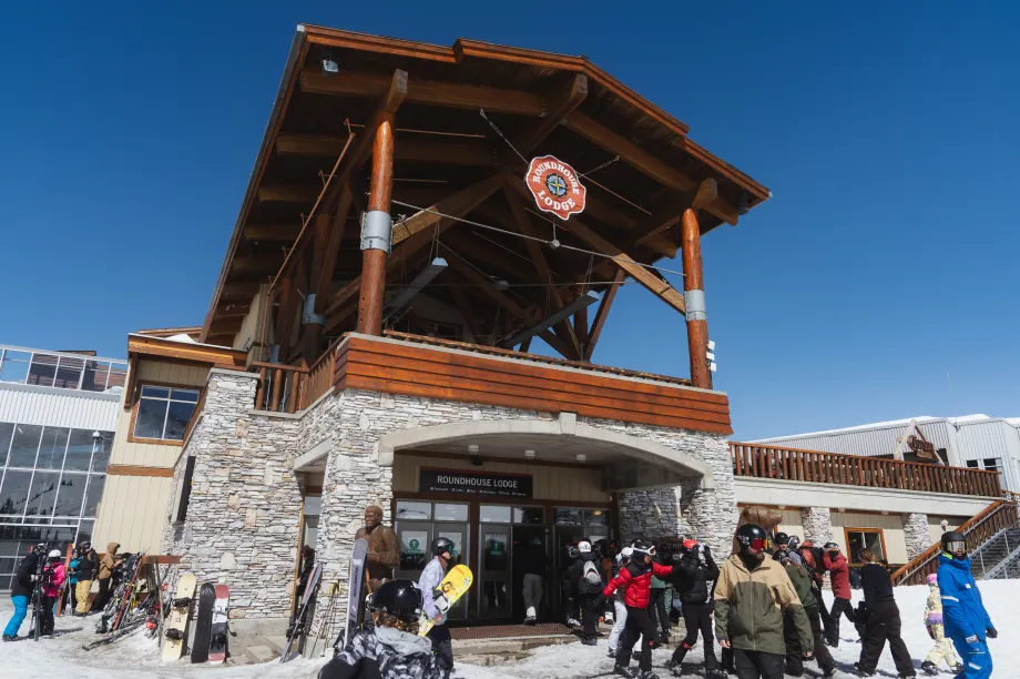 B&acirc;timent de chalet de ski avec une large entr&eacute;e de plain-pied et des portes doubles ouvertes. Des skieurs en tenue de ski se rassemblent &agrave; l'ext&eacute;rieur, sur une aire d'entr&eacute;e d&eacute;gag&eacute;e et plane avec une rambarde et de larges all&eacute;es menant au chalet.